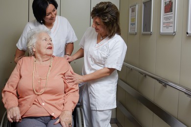 Zwei Pflegehelferinnen kümmern sich um eine Patientin im Rollstuhl