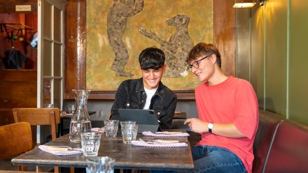 Zwei junge Männer, die ein JRK-Tandem bilden, sitzen an einem Tisch in einem Café und schauen gemeinsam auf ein iPad.